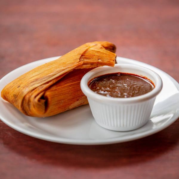A Noche Mexicana tamale wrapped in a corn husk, served with a side of dark mole sauce.