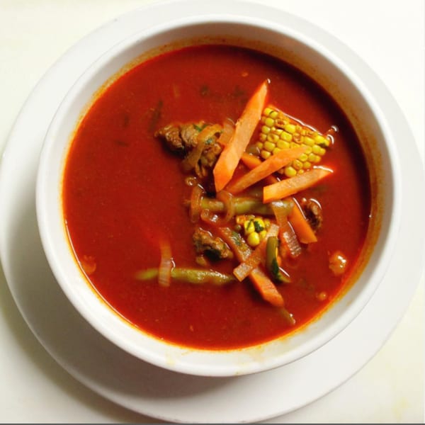 Noche Mexicana's red Mexican soup with corn, carrots, onions, and meat in a white bowl.