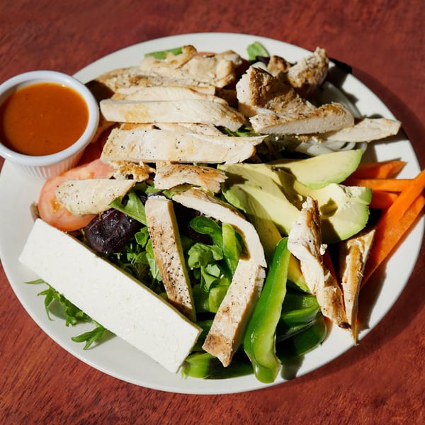 Grilled chicken salad with avocado, bell peppers, carrots, cheese, and a side of dressing.